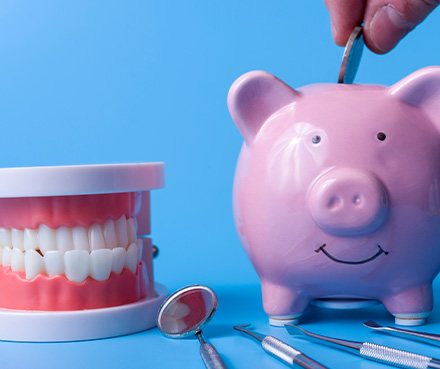 A hand putting a coin in a piggy bank next to dental tools and a jaw mockup