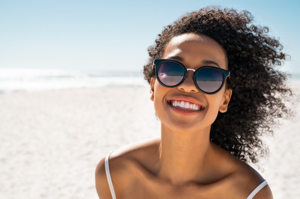 person with white teeth wearing sunglasses and smiling on beach